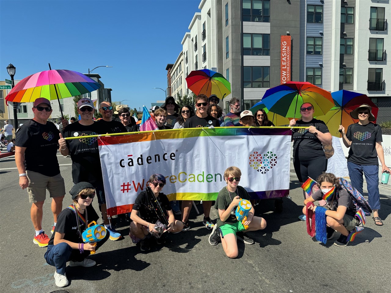 A group of Cadence employees and their family members smile with the company's Pride parade banner