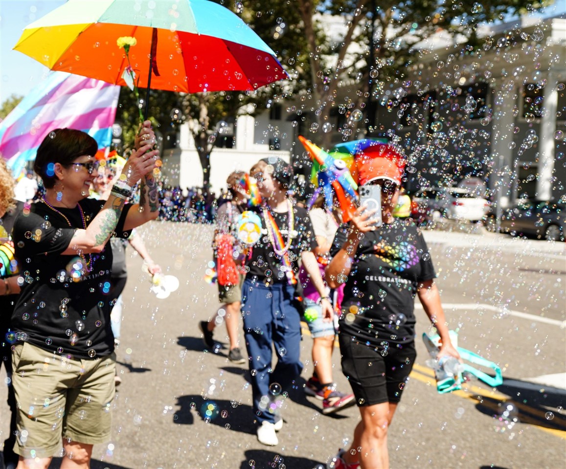 A woman is smiling under a colorful umbrella next to another woman taking pictures as bubbles float around them