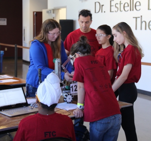 Four middle school kids and two adults in red shirts look at a robot that they built for a competition