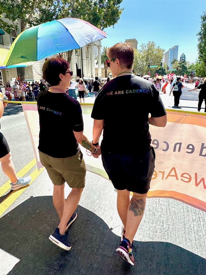 Two women are walking away from the camera and holding hands with t-shirts that read "We are Cadence."